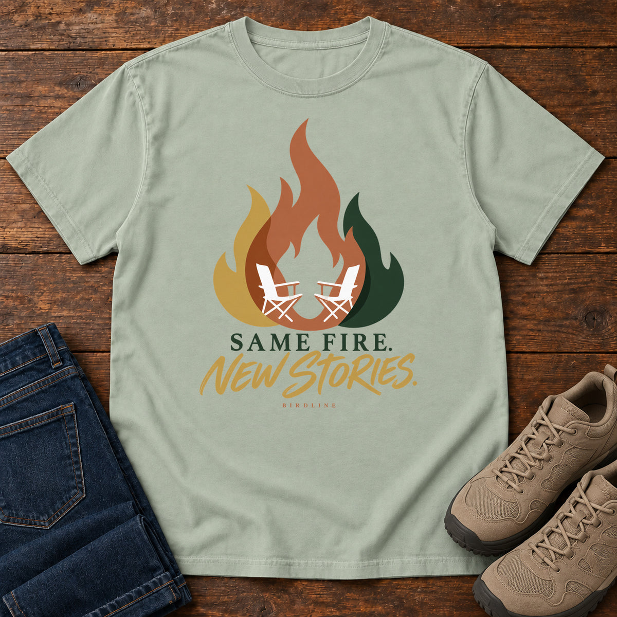 Bay Same Fire New Stories T-Shirt with a stylized orange, gold and green flame framing two white camp chairs; front flat lay on wood.