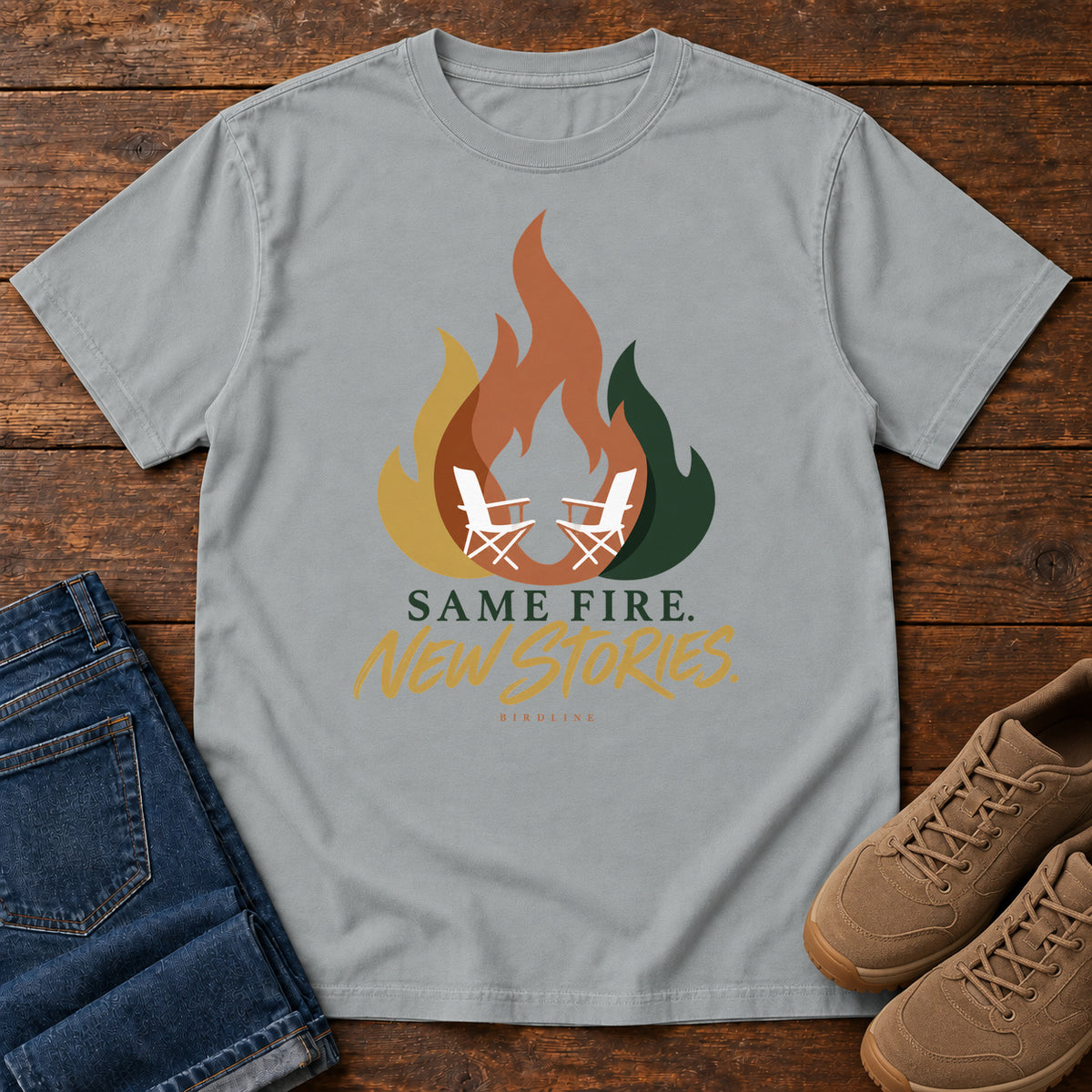 Granite Same Fire New Stories T-Shirt with a stylized orange, gold and green flame framing two white camp chairs; front flat lay on wood.