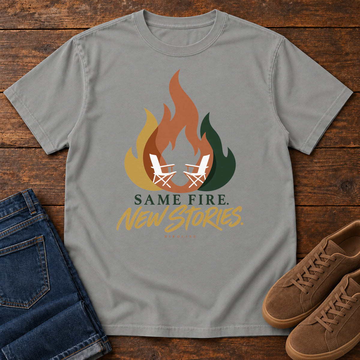 Grey Same Fire New Stories T-Shirt with a stylized orange, gold and green flame framing two white camp chairs; front flat lay on wood.