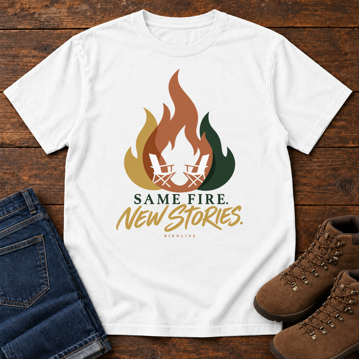 White Same Fire New Stories T-Shirt with a stylized orange, gold and green flame framing two white camp chairs; front flat lay on wood.