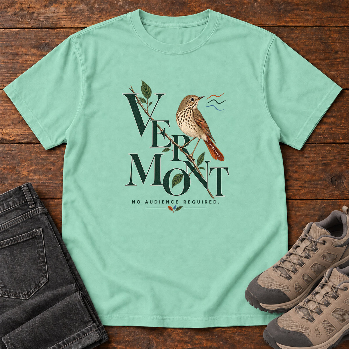 Island Reef Vermont No Audience Required Thrush T-Shirt with a brown speckle-breasted bird on a leafy branch among tall lettering; front flat lay on wood.