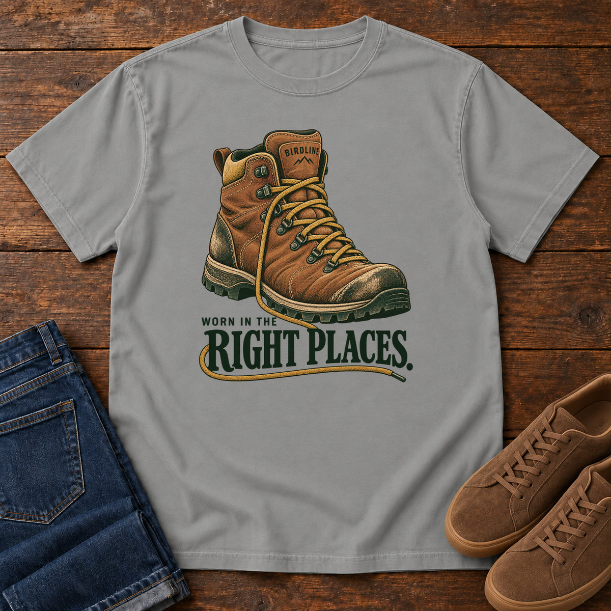 Worn in the Right Places T-Shirt in Grey, front laid flat on wood, featuring a brown lace-up hiking boot above dark green slogan lettering.