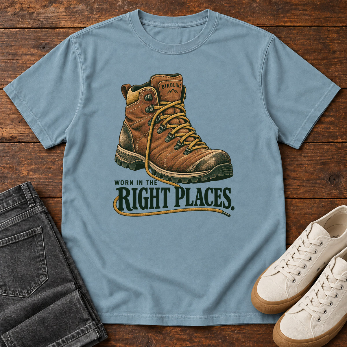Worn in the Right Places T-Shirt in Ice Blue, front laid flat on wood, featuring a brown lace-up hiking boot above dark green slogan lettering.