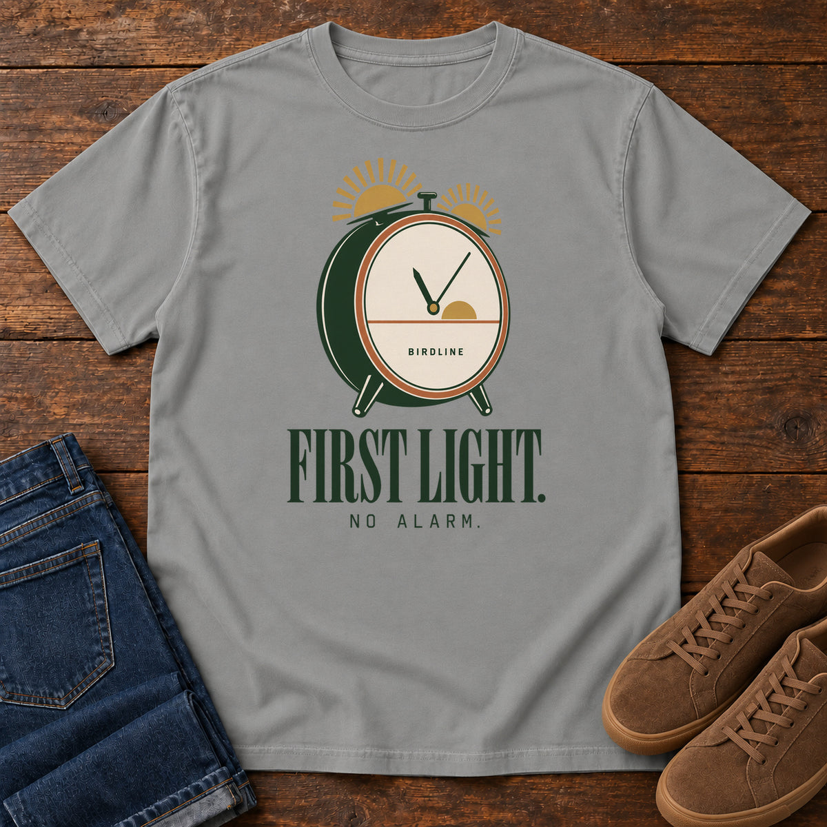 Grey First Light No Alarm T-Shirt with a green-and-cream clock with gold sunrise shapes above First Light No Alarm lettering, front flat lay on wood.