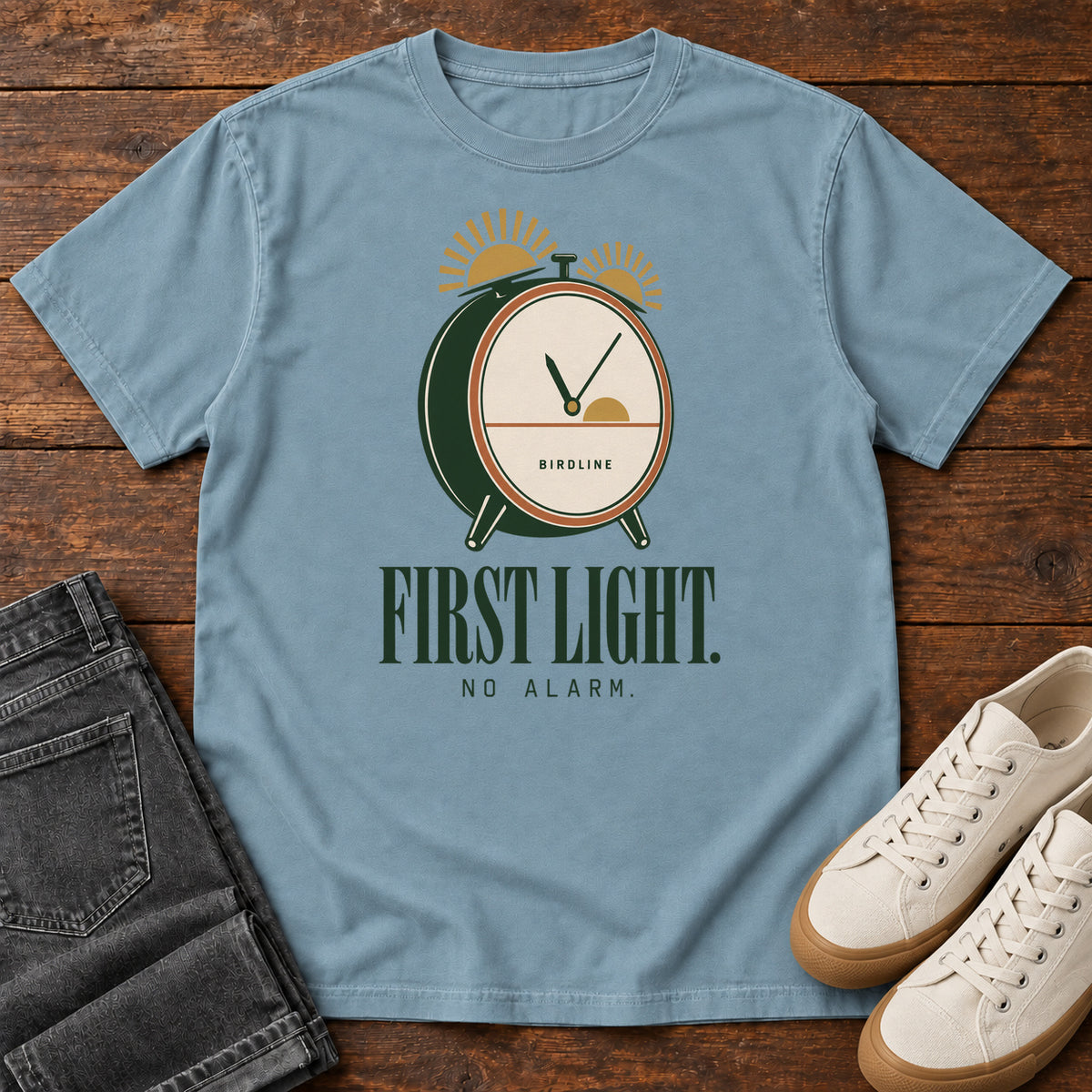 Ice Blue First Light No Alarm T-Shirt with a green-and-cream clock with gold sunrise shapes above First Light No Alarm lettering, front flat lay on wood.