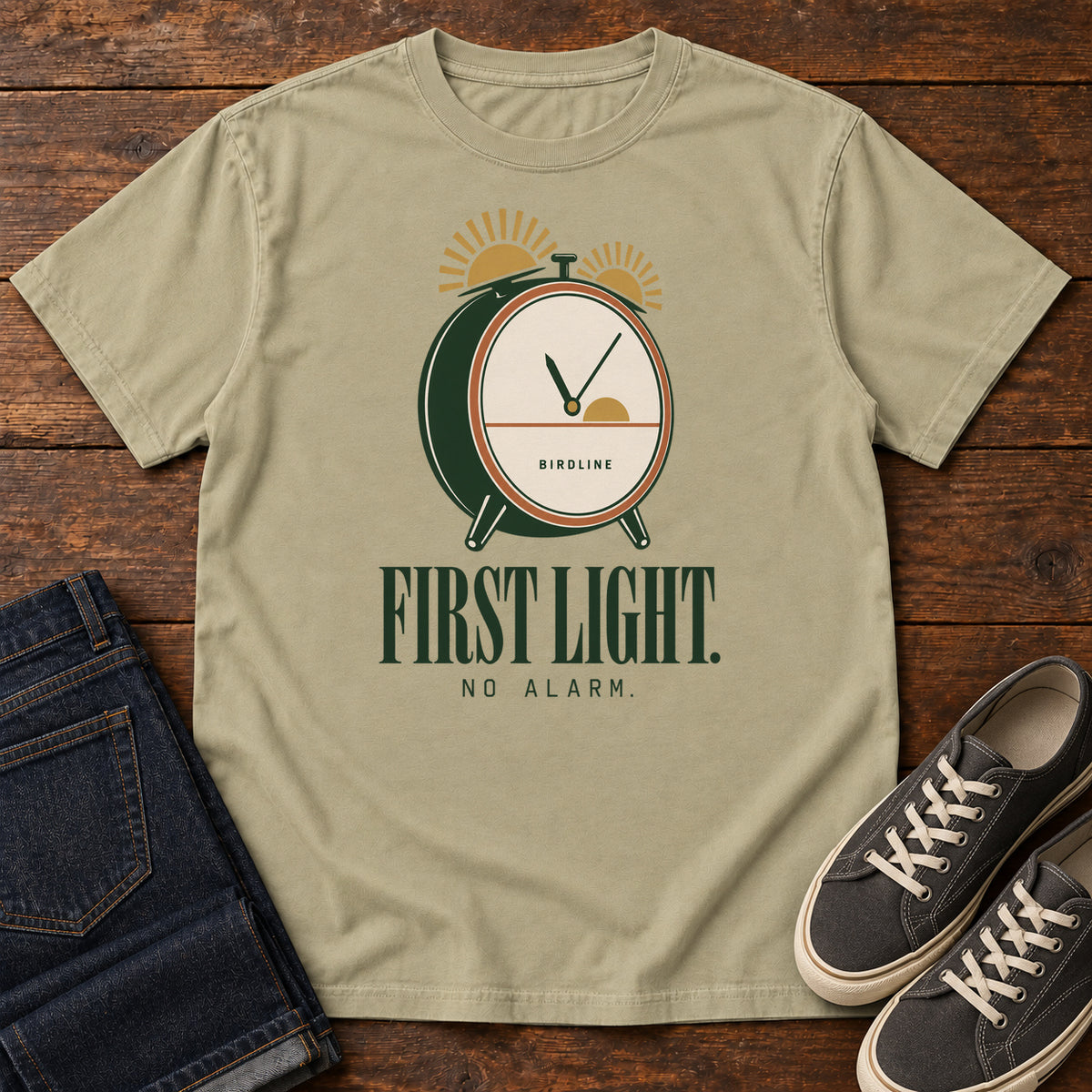 Khaki First Light No Alarm T-Shirt with a green-and-cream clock with gold sunrise shapes above First Light No Alarm lettering, front flat lay on wood.
