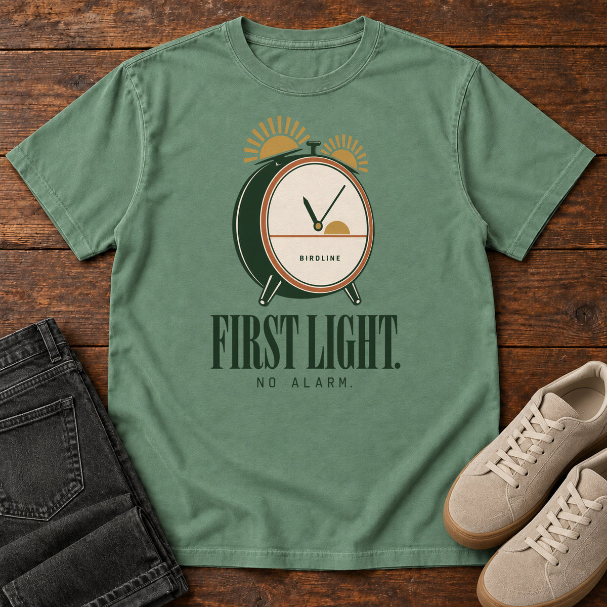 Light Green First Light No Alarm T-Shirt with a green-and-cream clock with gold sunrise shapes above First Light No Alarm lettering, front flat lay on wood.