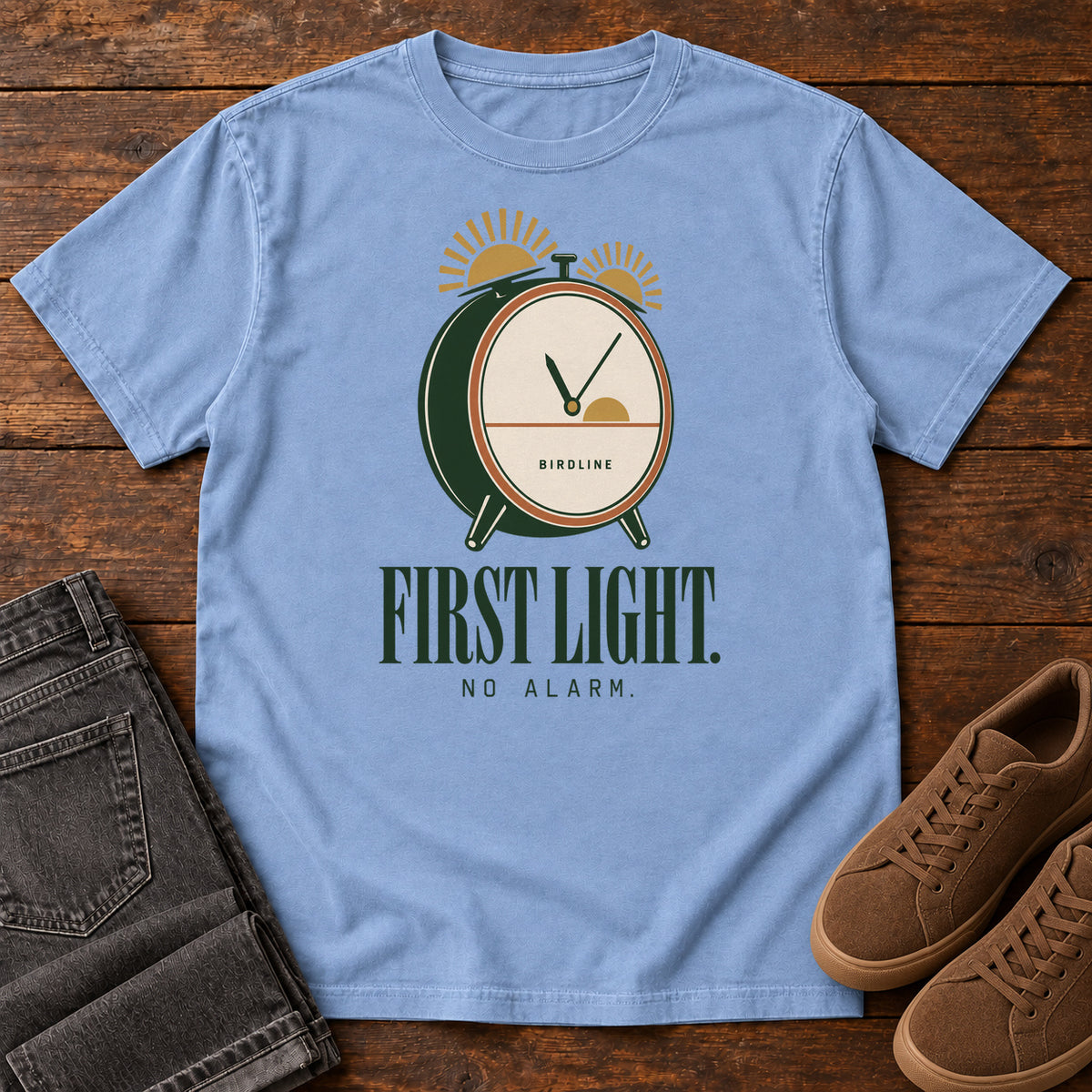 Washed Denim First Light No Alarm T-Shirt with a green-and-cream clock with gold sunrise shapes above First Light No Alarm lettering, front flat lay on wood.