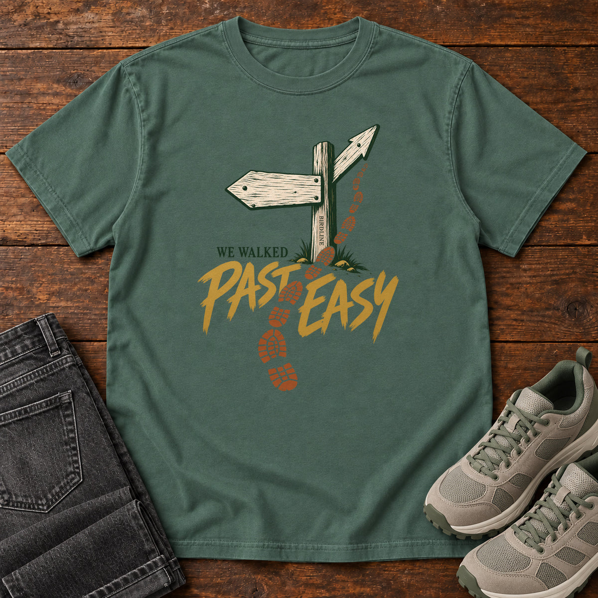 Blue Spruce We Walked Past Easy T-Shirt with a wooden trail sign, orange boot prints and golden script, front flat lay on wood.
