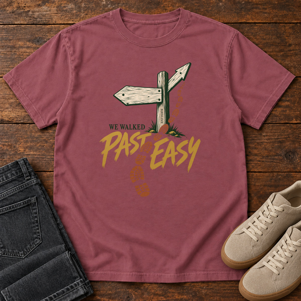 Brick We Walked Past Easy T-Shirt with a wooden trail sign, orange boot prints and golden script, front flat lay on wood.