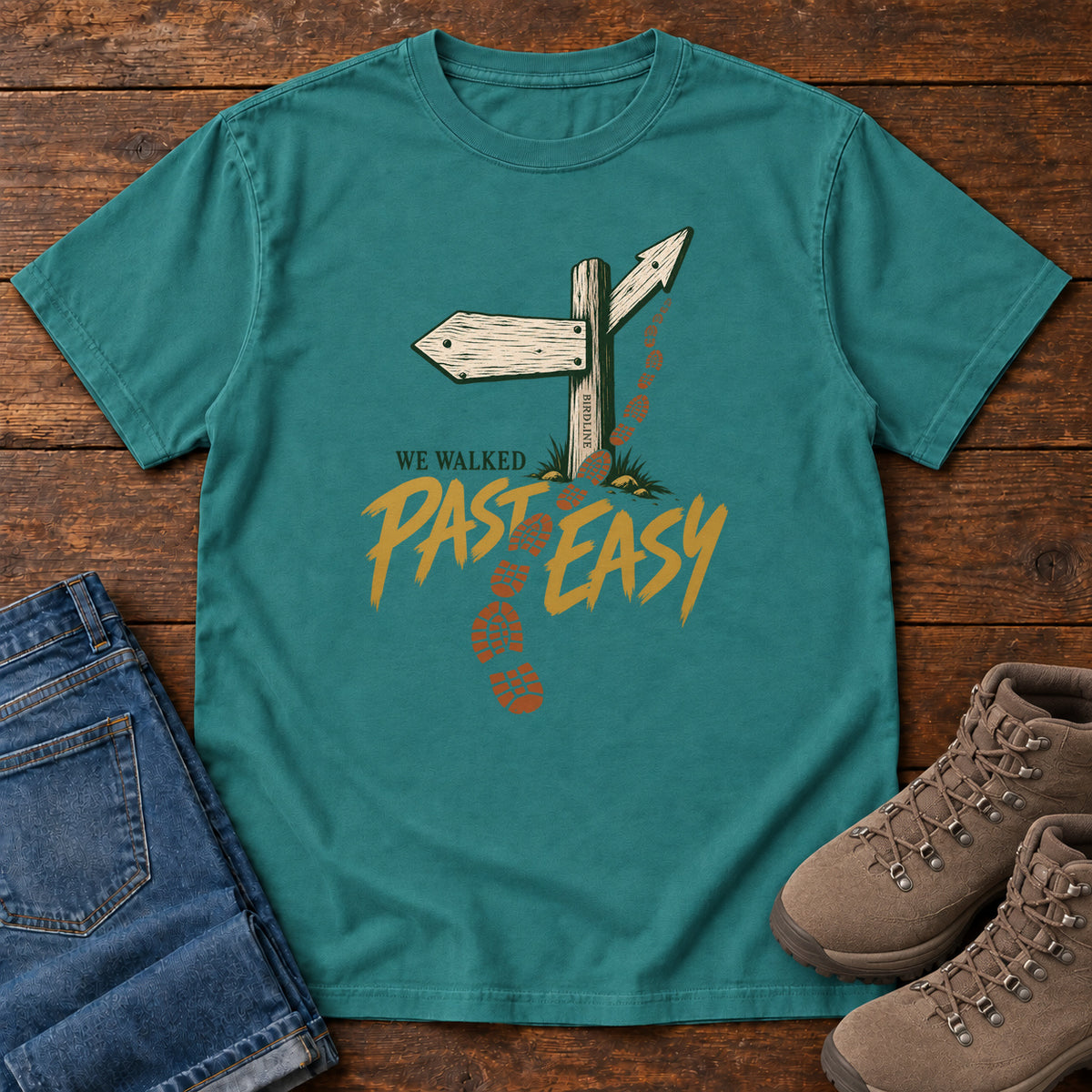 Emerald We Walked Past Easy T-Shirt with a wooden trail sign, orange boot prints and golden script, front flat lay on wood.
