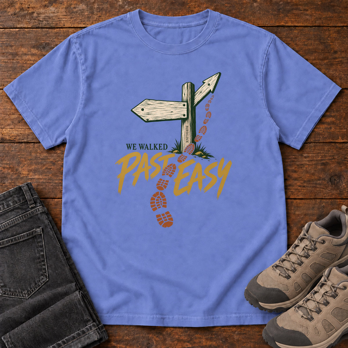 Flo Blue We Walked Past Easy T-Shirt with a wooden trail sign, orange boot prints and golden script, front flat lay on wood.