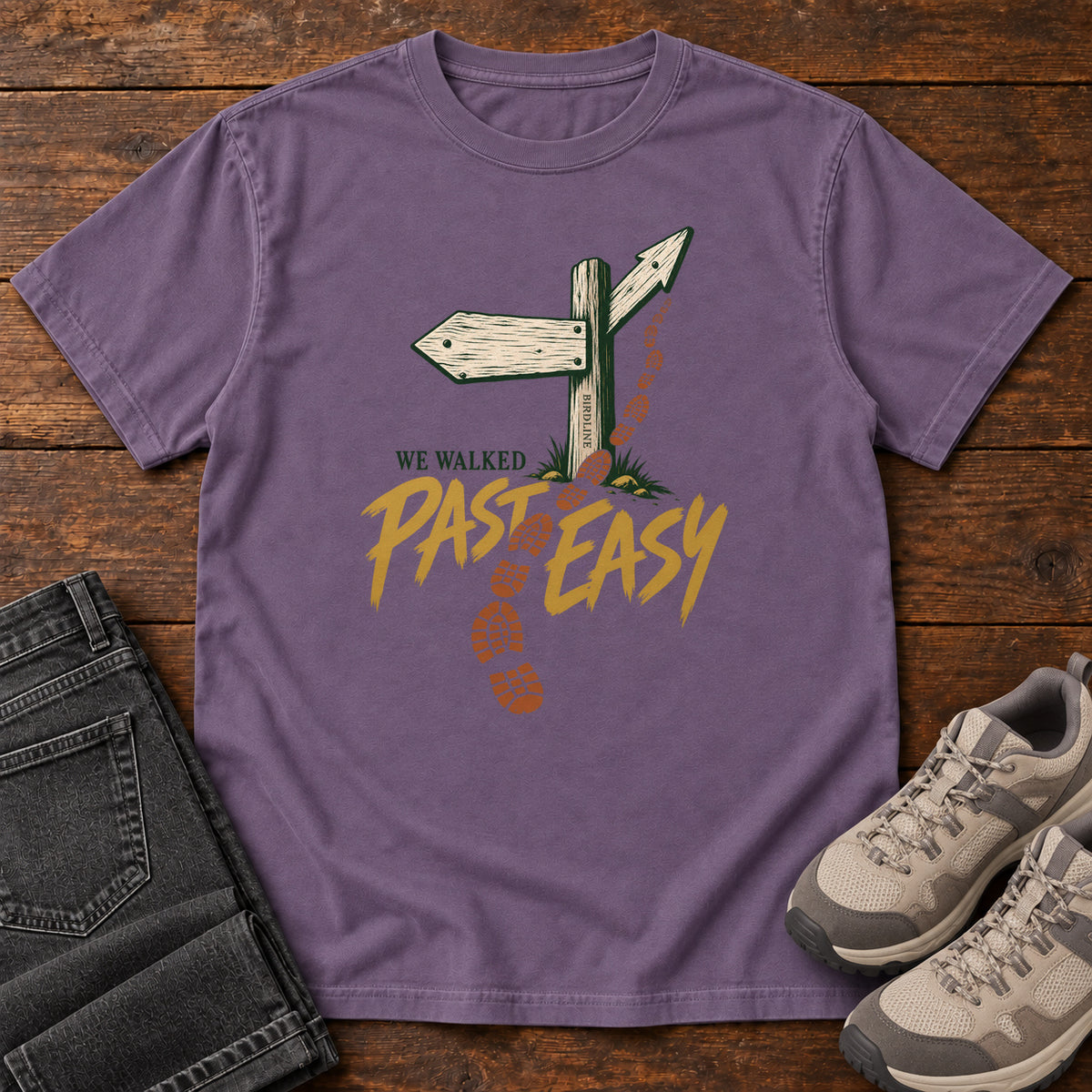 Grape We Walked Past Easy T-Shirt with a wooden trail sign, orange boot prints and golden script, front flat lay on wood.
