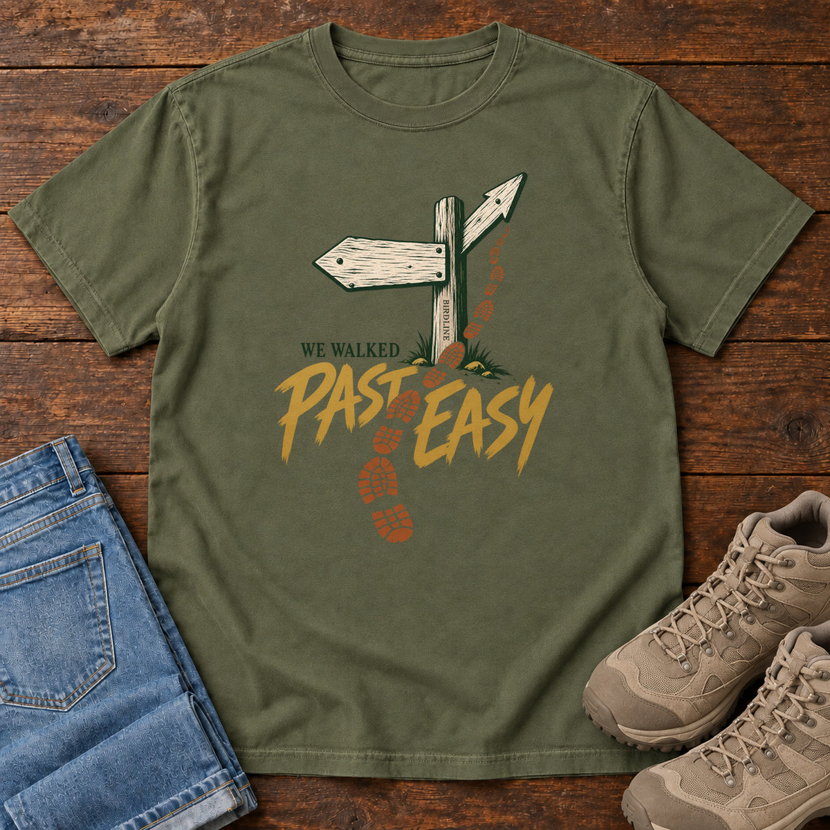 Hemp We Walked Past Easy T-Shirt with a wooden trail sign, orange boot prints and golden script, front flat lay on wood.