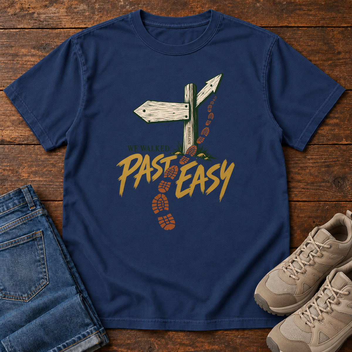 True Navy We Walked Past Easy T-Shirt with a wooden trail sign, orange boot prints and golden script, front flat lay on wood.