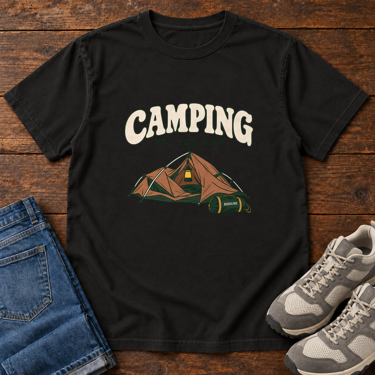 Black Camping Was the Plan T-Shirt with an orange tent with a hanging lantern and green gear bag; front flat lay on wood.