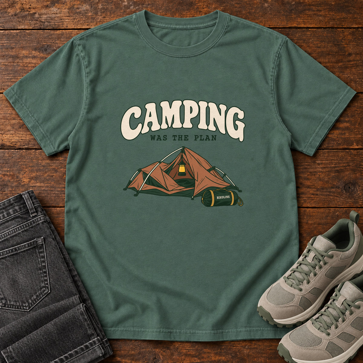 Blue Spruce Camping Was the Plan T-Shirt with an orange tent with a hanging lantern and green gear bag; front flat lay on wood.