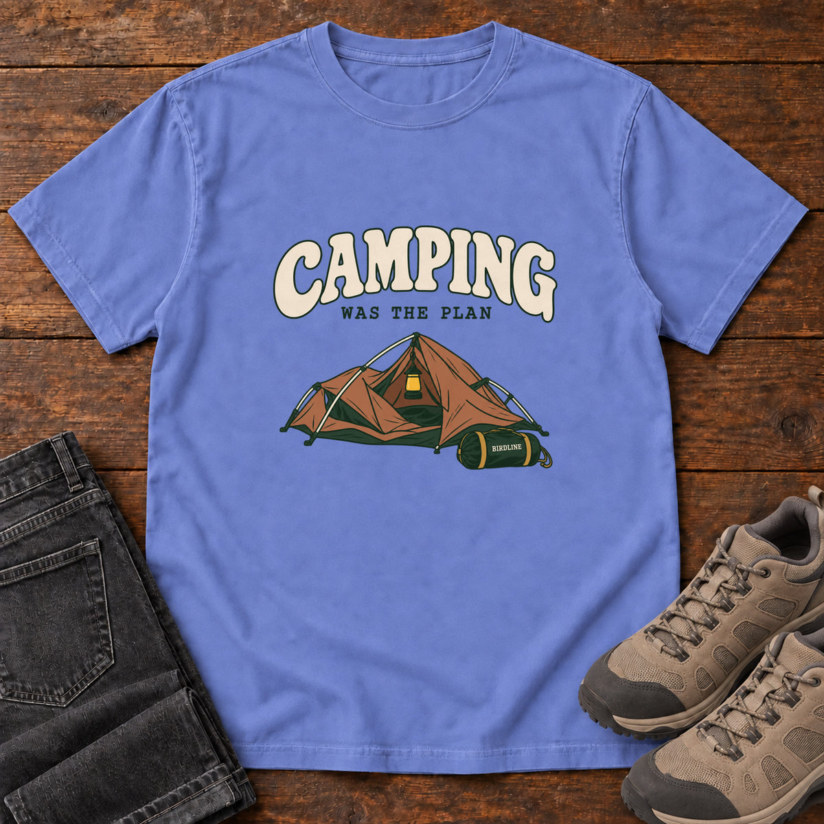 Flo Blue Camping Was the Plan T-Shirt with an orange tent with a hanging lantern and green gear bag; front flat lay on wood.
