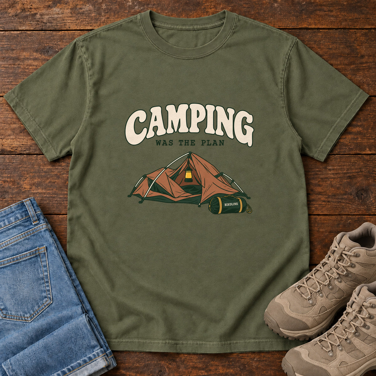 Hemp Camping Was the Plan T-Shirt with an orange tent with a hanging lantern and green gear bag; front flat lay on wood.