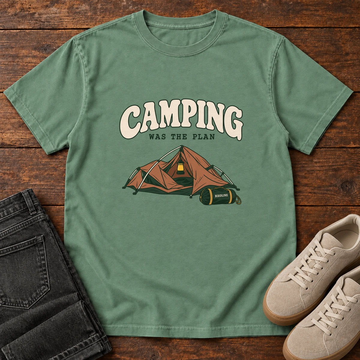 Light Green Camping Was the Plan T-Shirt with an orange tent with a hanging lantern and green gear bag; front flat lay on wood.