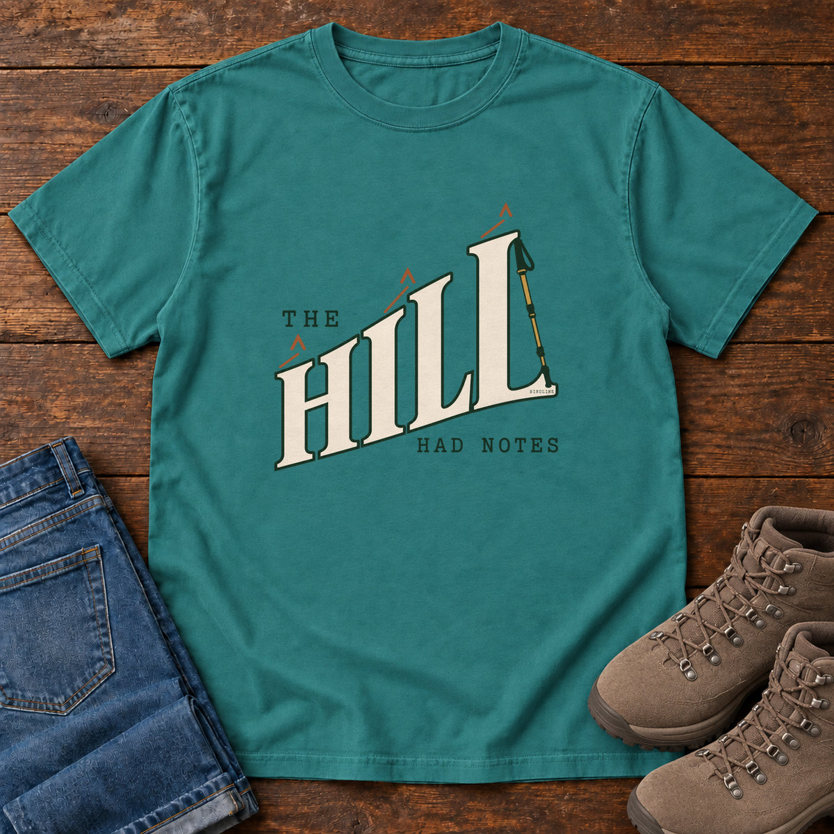 The Hill Had Notes T-Shirt in Emerald, front laid flat on wood, featuring ascending cream HILL lettering with small orange chevrons and a trekking pole.