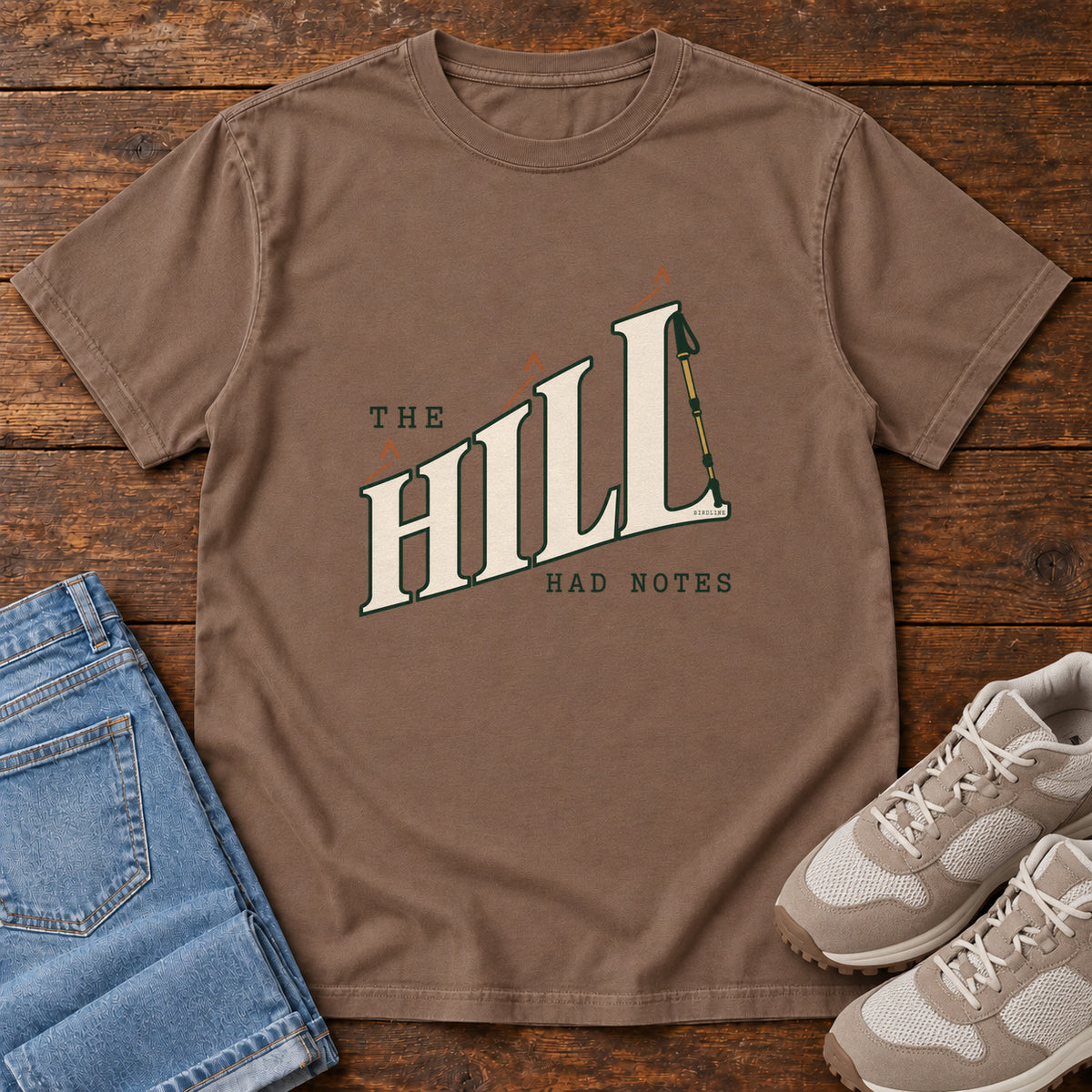 The Hill Had Notes T-Shirt in Espresso, front laid flat on wood, featuring ascending cream HILL lettering with small orange chevrons and a trekking pole.
