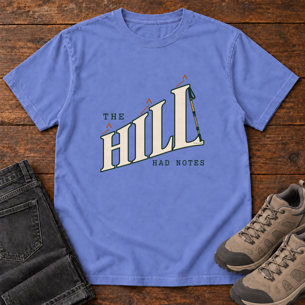 The Hill Had Notes T-Shirt in Flo Blue, front laid flat on wood, featuring ascending cream HILL lettering with small orange chevrons and a trekking pole.