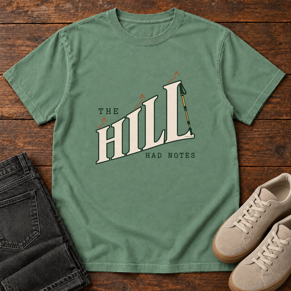 The Hill Had Notes T-Shirt in Light Green, front laid flat on wood, featuring ascending cream HILL lettering with small orange chevrons and a trekking pole.
