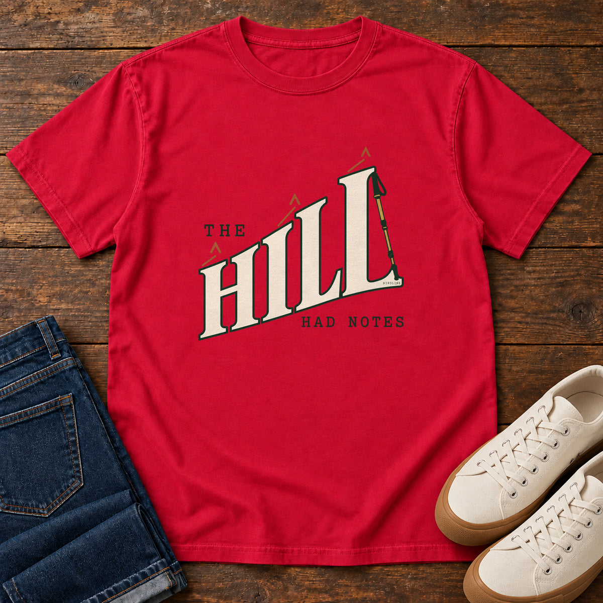 The Hill Had Notes T-Shirt in Red, front laid flat on wood, featuring ascending cream HILL lettering with small orange chevrons and a trekking pole.