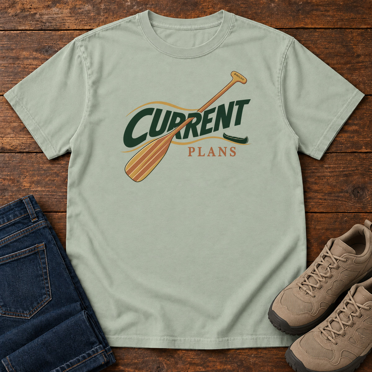 Bay Current Plans T-Shirt with a diagonal wooden paddle crossing green and gold Current Plans lettering; front flat lay on wood.