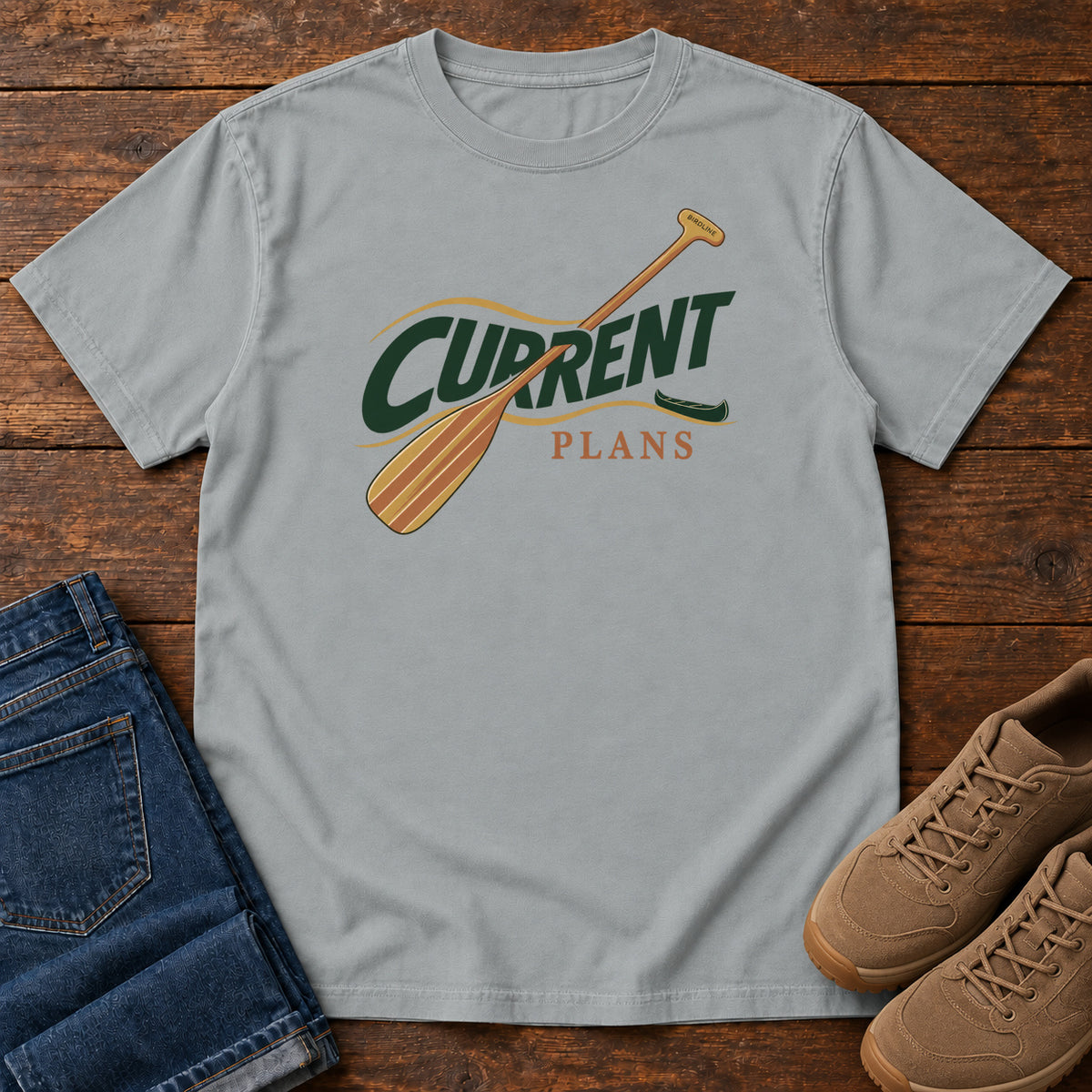 Granite Current Plans T-Shirt with a diagonal wooden paddle crossing green and gold Current Plans lettering; front flat lay on wood.