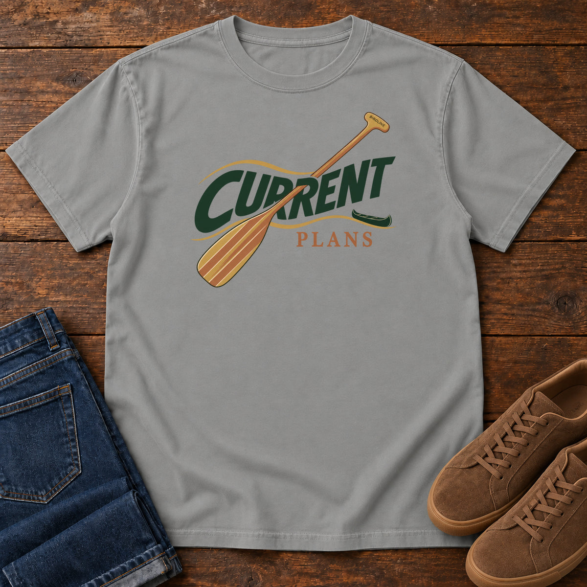 Grey Current Plans T-Shirt with a diagonal wooden paddle crossing green and gold Current Plans lettering; front flat lay on wood.