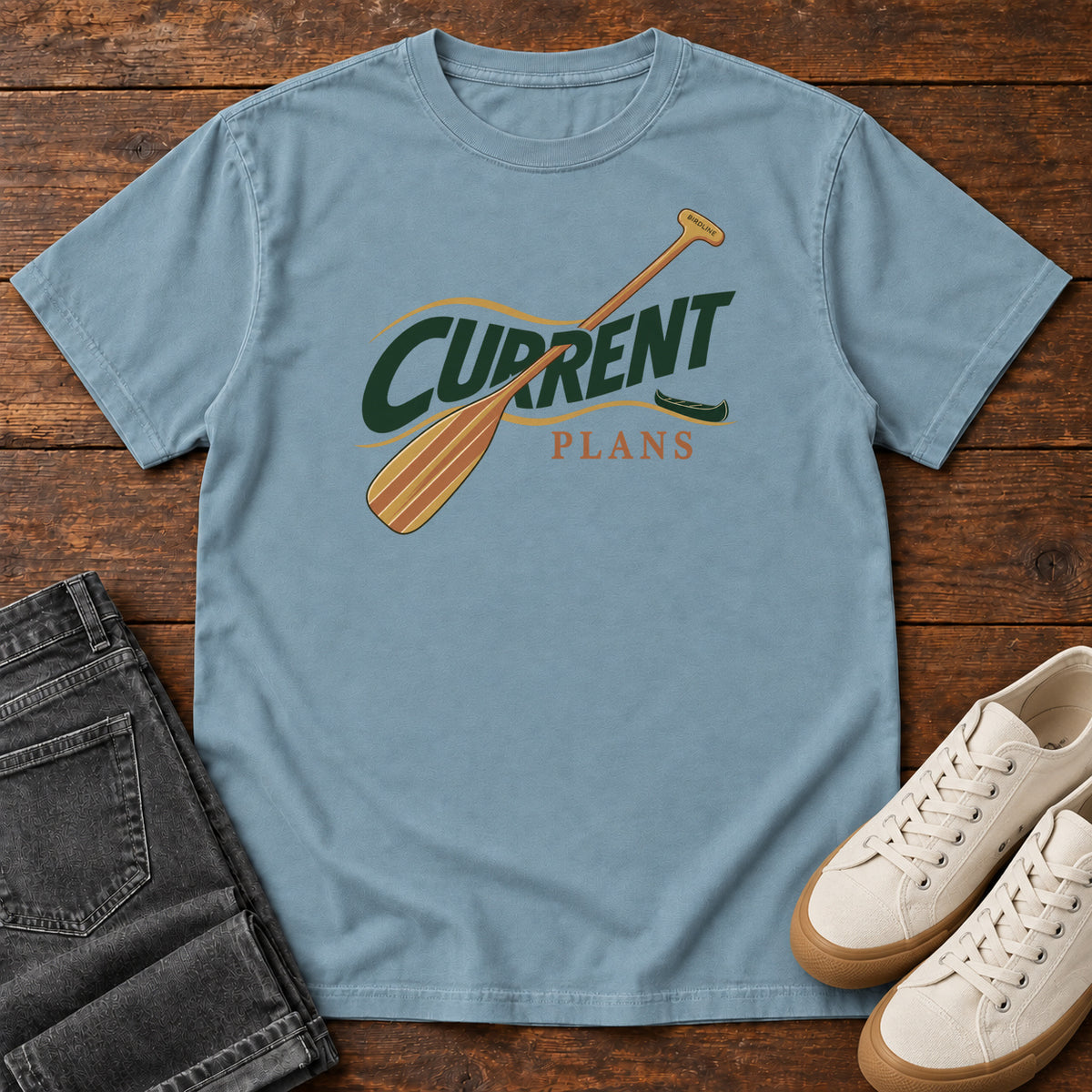 Ice Blue Current Plans T-Shirt with a diagonal wooden paddle crossing green and gold Current Plans lettering; front flat lay on wood.