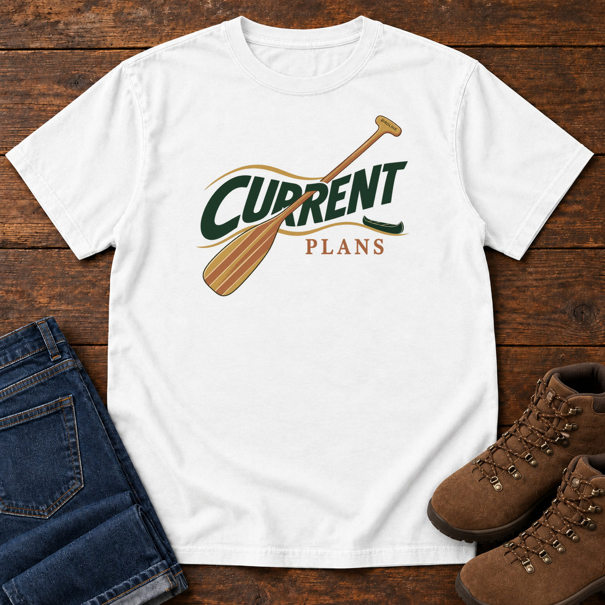 White Current Plans T-Shirt with a diagonal wooden paddle crossing green and gold Current Plans lettering; front flat lay on wood.