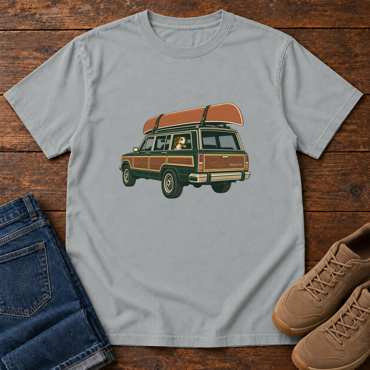 Granite Canoe Wagon and Dog T-Shirt with a green wood-paneled wagon, roof-mounted canoe and dog in the window, front flat lay on wood.