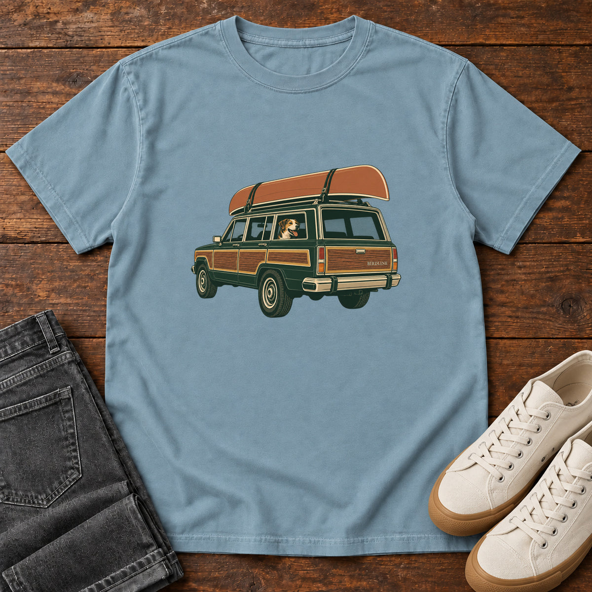 Ice Blue Canoe Wagon and Dog T-Shirt with a green wood-paneled wagon, roof-mounted canoe and dog in the window, front flat lay on wood.