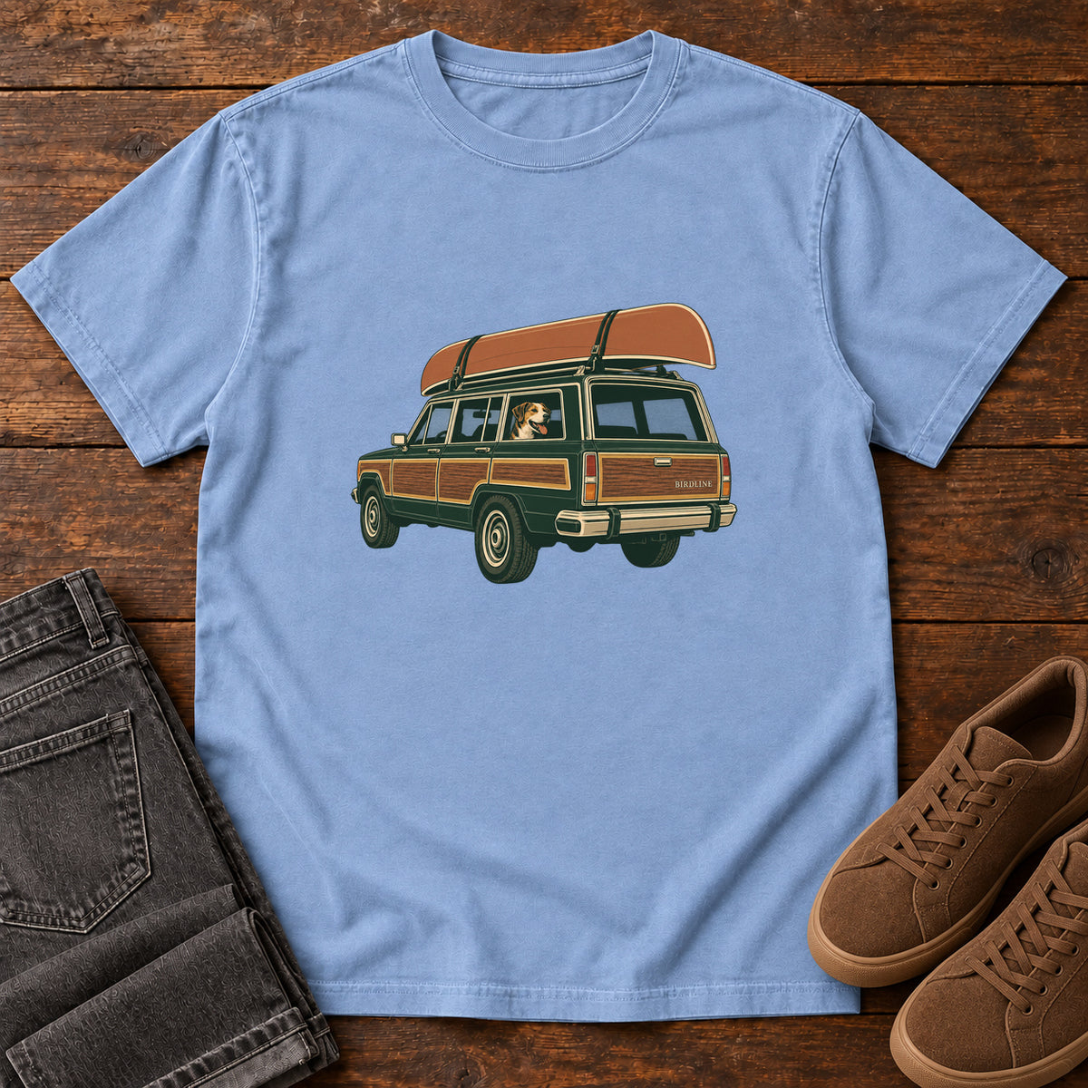Washed Denim Canoe Wagon and Dog T-Shirt with a green wood-paneled wagon, roof-mounted canoe and dog in the window, front flat lay on wood.