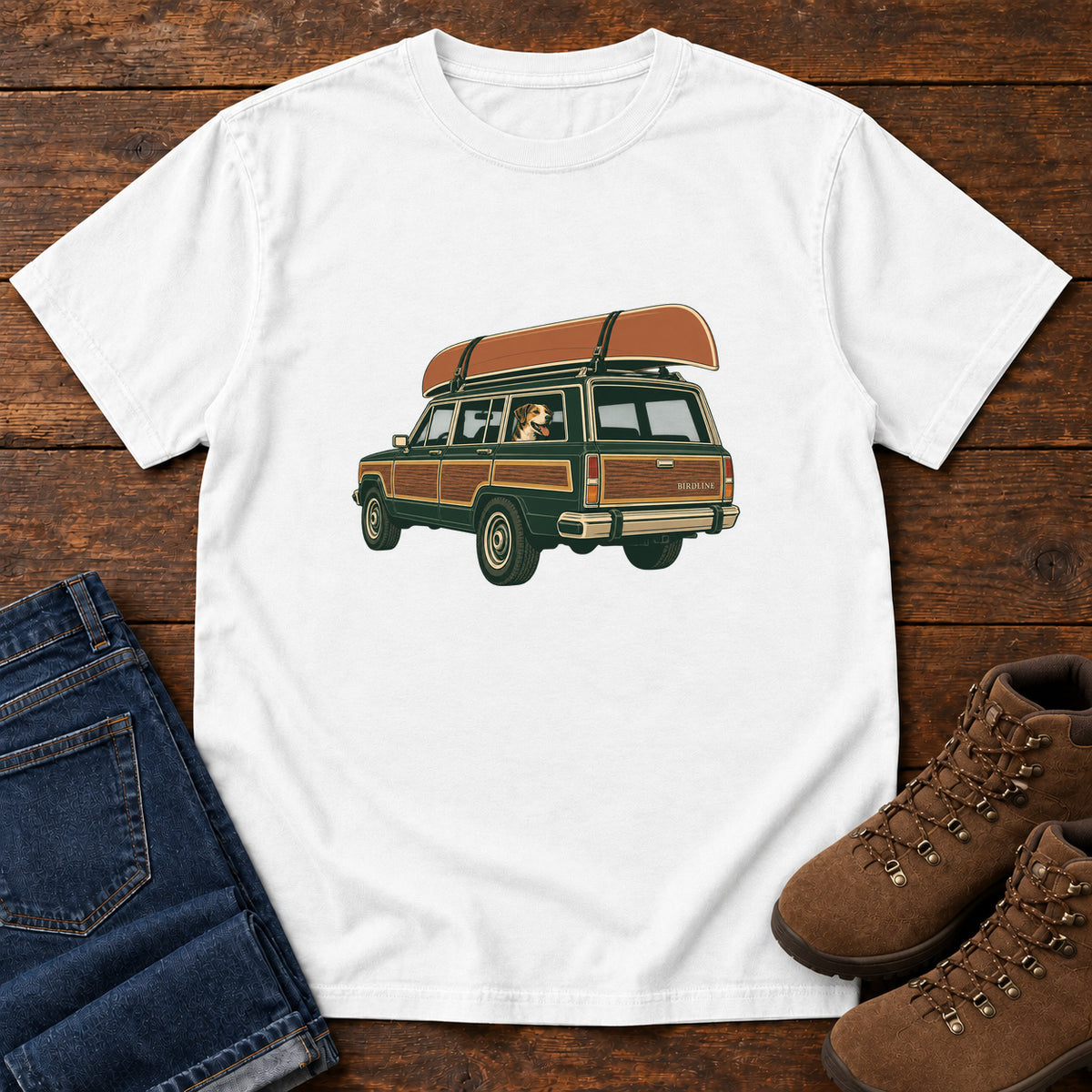 White Canoe Wagon and Dog T-Shirt with a green wood-paneled wagon, roof-mounted canoe and dog in the window, front flat lay on wood.
