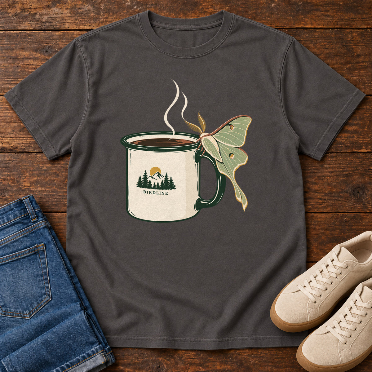 Graphite Coffee and Luna Moth T-Shirt with a steaming cream camp mug with green trim and a pale green long-tailed moth; front flat lay on wood.