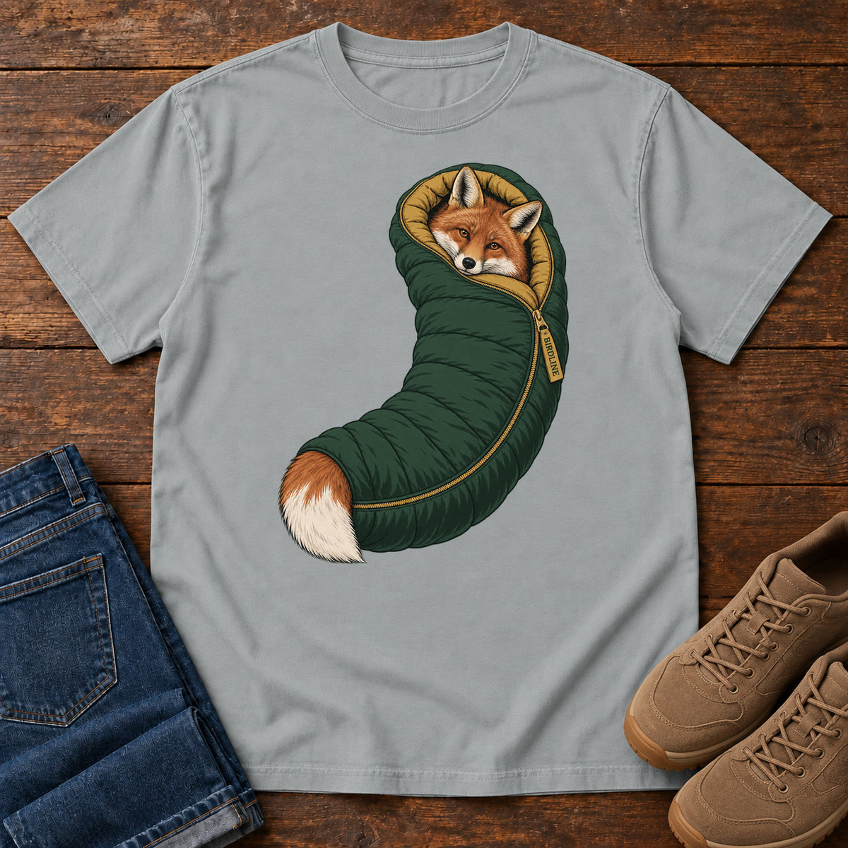 Granite Fox in a Sleeping Bag T-Shirt with an orange fox tucked into a green sleeping bag with its white-tipped tail showing; front flat lay on wood.