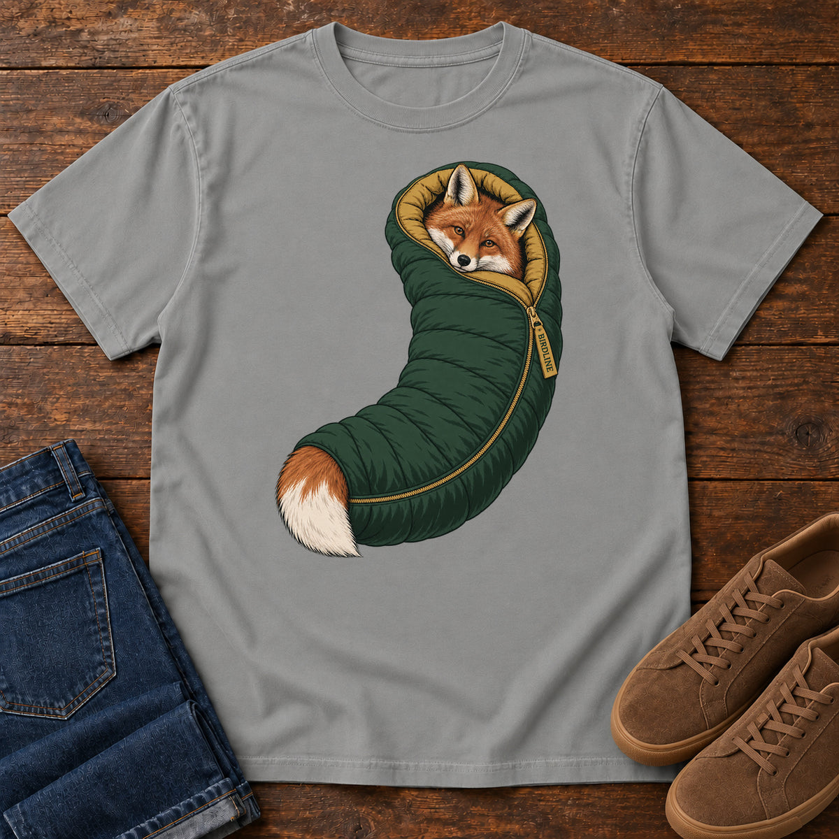 Grey Fox in a Sleeping Bag T-Shirt with an orange fox tucked into a green sleeping bag with its white-tipped tail showing; front flat lay on wood.