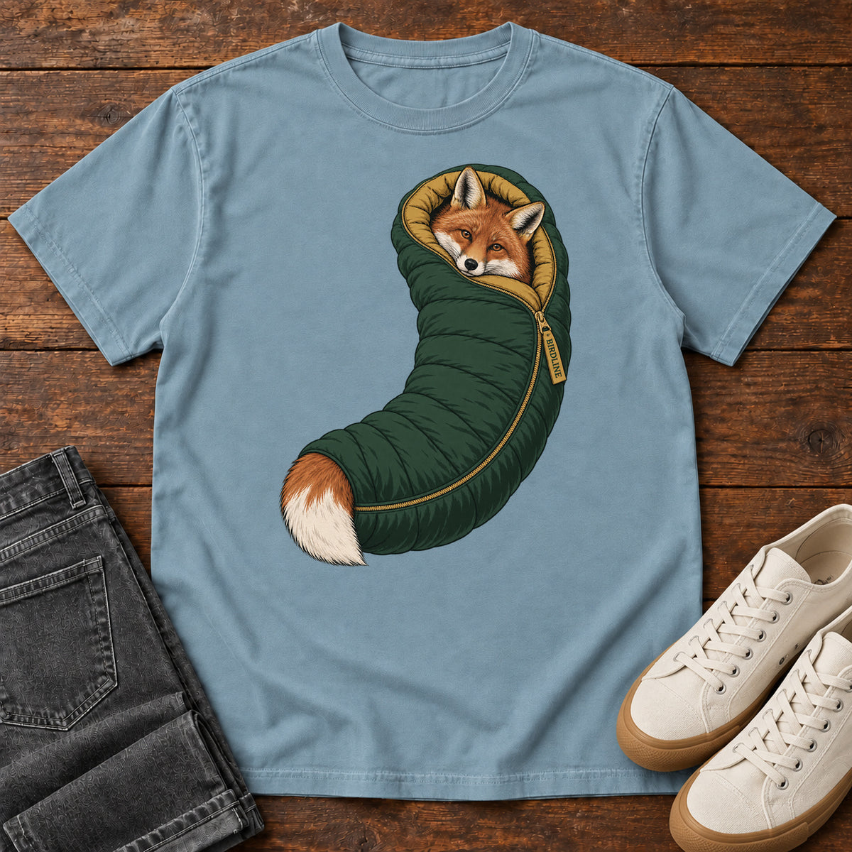 Ice Blue Fox in a Sleeping Bag T-Shirt with an orange fox tucked into a green sleeping bag with its white-tipped tail showing; front flat lay on wood.