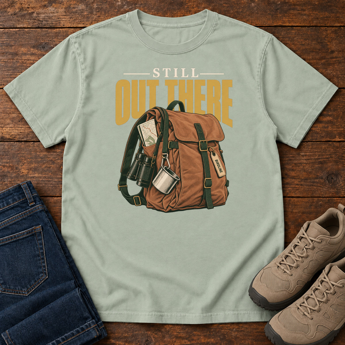 Bay Still Out There T-Shirt with a brown hiking backpack with binoculars, a mug and gold lettering, front flat lay on wood.