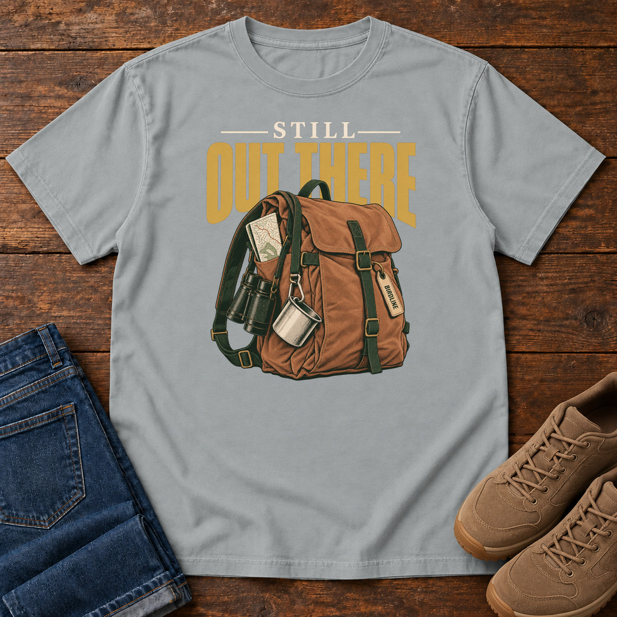 Granite Still Out There T-Shirt with a brown hiking backpack with binoculars, a mug and gold lettering, front flat lay on wood.