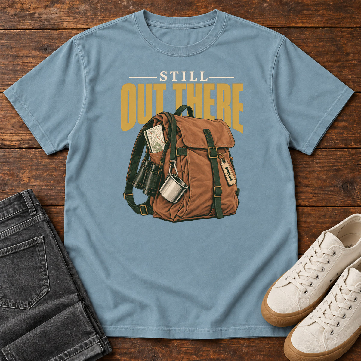 Ice Blue Still Out There T-Shirt with a brown hiking backpack with binoculars, a mug and gold lettering, front flat lay on wood.