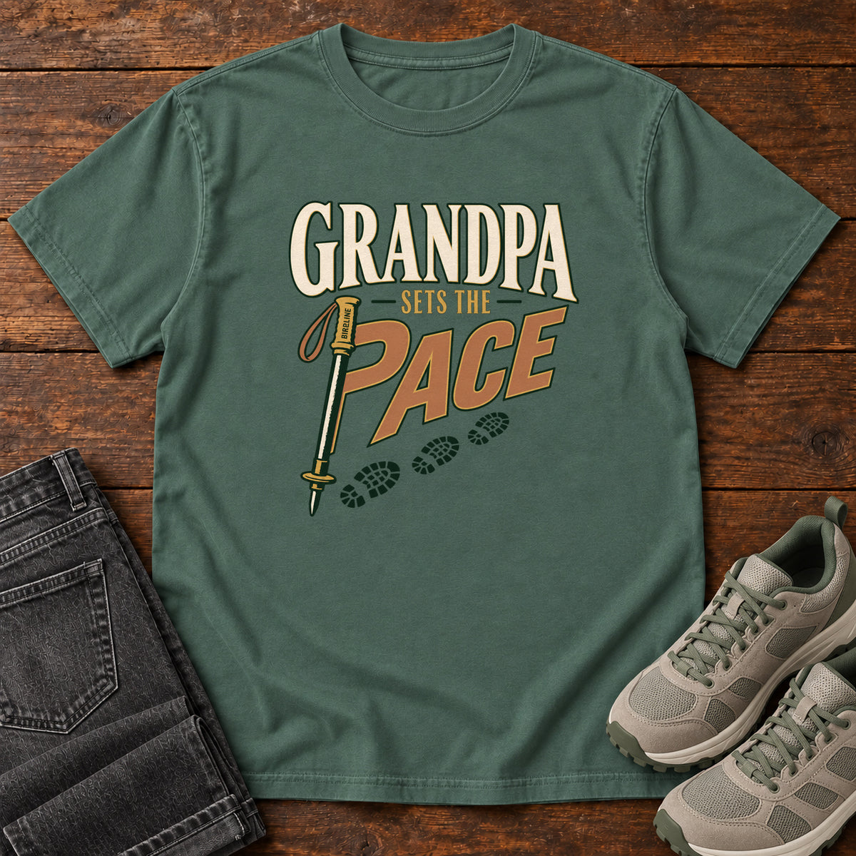 Grandpa Sets the Pace T-Shirt in Blue Spruce, front laid flat on wood, featuring cream-and-orange slogan lettering beside a trekking pole and a trail of boot prints.
