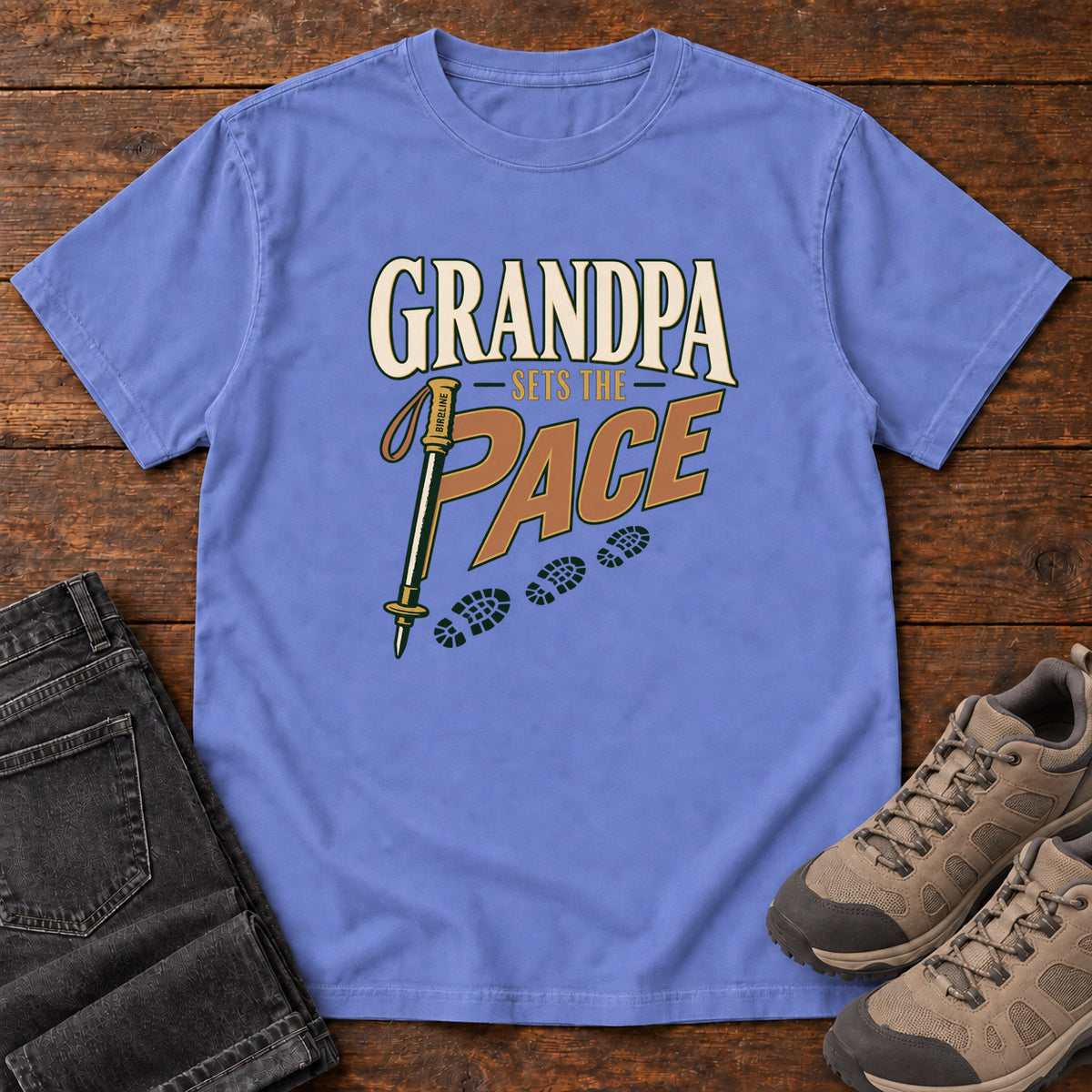 Grandpa Sets the Pace T-Shirt in Flo Blue, front laid flat on wood, featuring cream-and-orange slogan lettering beside a trekking pole and a trail of boot prints.