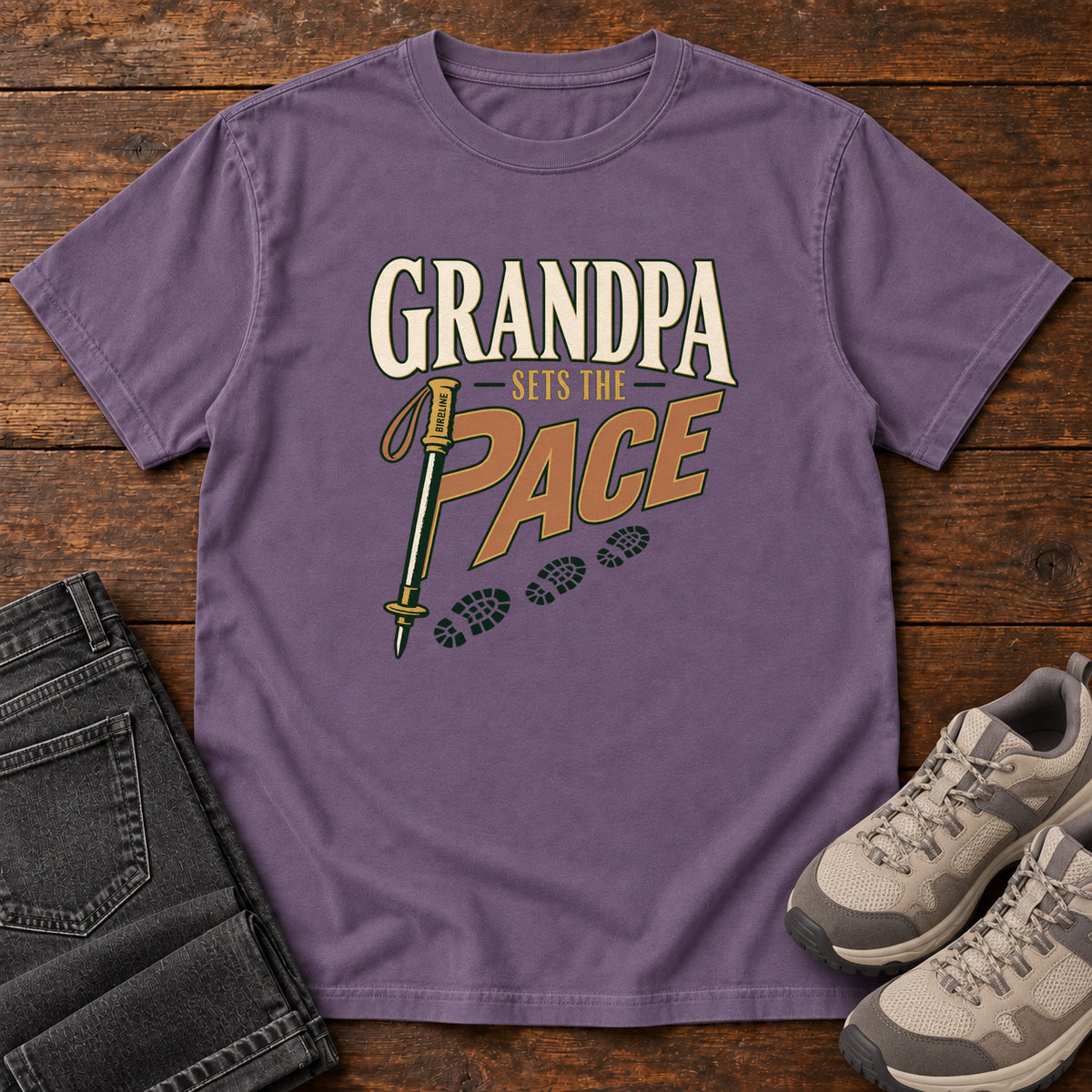 Grandpa Sets the Pace T-Shirt in Grape, front laid flat on wood, featuring cream-and-orange slogan lettering beside a trekking pole and a trail of boot prints.