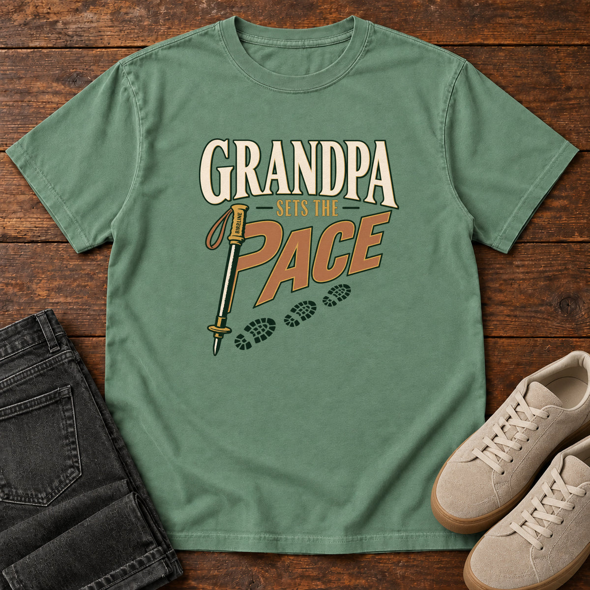 Grandpa Sets the Pace T-Shirt in Light Green, front laid flat on wood, featuring cream-and-orange slogan lettering beside a trekking pole and a trail of boot prints.