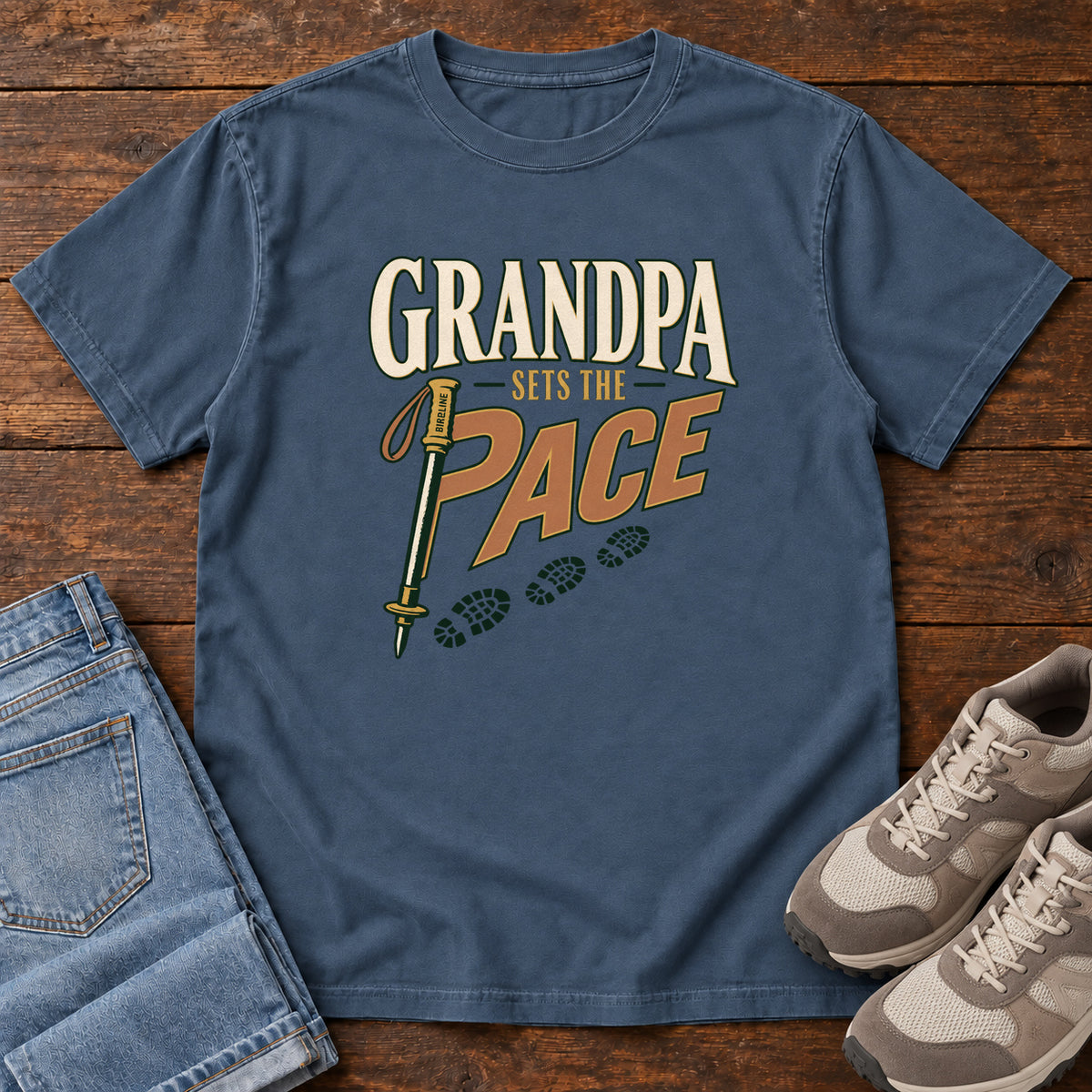 Grandpa Sets the Pace T-Shirt in Midnight, front laid flat on wood, featuring cream-and-orange slogan lettering beside a trekking pole and a trail of boot prints.